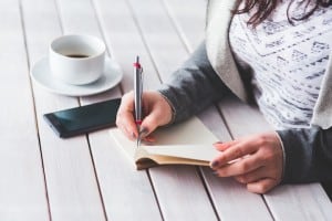 Woman writing in notebook with smartphone and coffee next to her