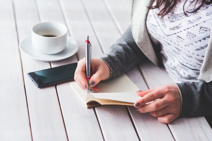 Woman writing in notebook with smartphone and coffee next to her
